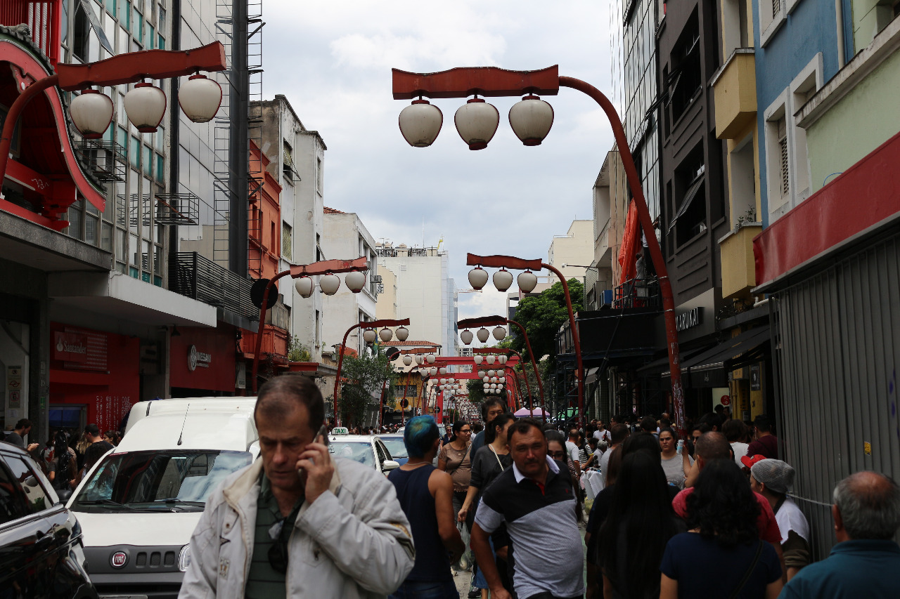 Fotografia colorida mostra rua Galvão Beno, no bairro da Liberdade repleta de pessoas.  É possível avistar diversos postes de iluminação no estilo japonês